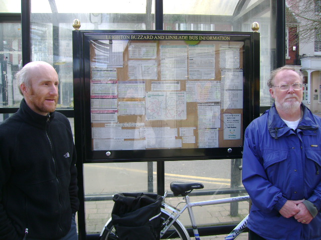 Two members of the group stand in front of the bus information at the bus stop in the town centre.